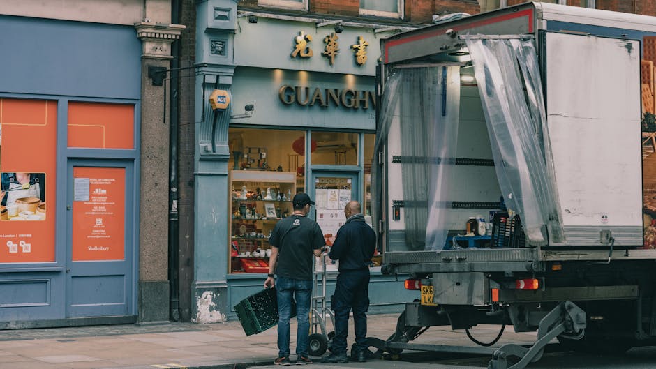 Two men with dark hair, one wearing a black baseball cap and the other with a bald head, are standing outside a shop with a blue facade and Chinese characters above the window, which displays ceramic dishes and decorative items inside. The men are engaged in conversation near a large, open-moving truck parked on the pavement, with clear plastic wrapping draped over the truck's rear cargo area. One of the men is holding a small trolley, and a green plastic crate is on the ground nearby. The scene is set in an urban environment during daylight, with the truck likely being used for furniture transport and packing, relevant to home relocation or last-minute removals facilitated by South Kensington Removals.
