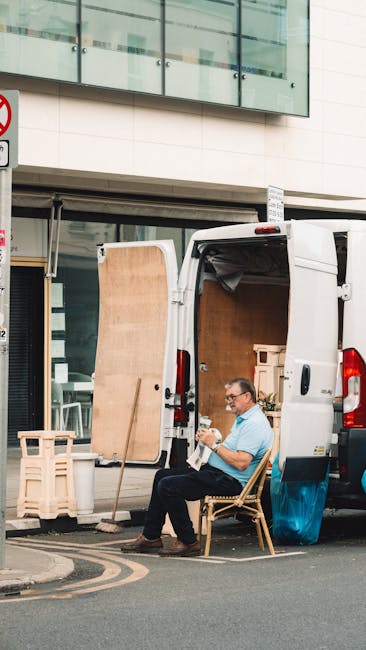 A man seated on a wooden chair is resting outside a white commercial van parked on the street in South Kensington, during a home relocation process. The van's rear doors are open, revealing a large, flat wooden panel leaning against the vehicle and various packing materials. Inside the van, visible cardboard boxes and wrapped furniture items indicate an ongoing packing and moving operation. The man, dressed casually in a light blue shirt and dark trousers, appears to be taking a break from loading or unloading furniture with the assistance of equipment such as straps or trolleys, though these are not visible in this frame. The surrounding environment includes a modern building with glass windows and a reflective façade, and a street sign is partially visible at the left edge of the image. This scene represents the logistics of furniture transport and packing that are typical in last-minute or delayed house removals handled by South Kensington Removals, reflecting professional moving services and careful handling of belongings for an efficient home relocation.