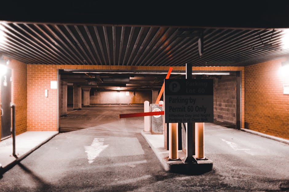 Inside a multi-storey underground parking garage with a brick wall exterior visible at the entrance. The area is dimly lit with overhead lighting. In the foreground, there is a parking barrier gate with an orange arm raised, and a sign indicating parking restrictions for permit holders in parking lot 60. Several concrete wheel stops are visible on the asphalt floor, which features directional white arrows painted on the surface. The parking spaces are empty, leading into a covered loading area where furniture, cardboard boxes, and moving supplies are typically transported during a home relocation. South Kensington Removals occasionally operate within such facilities to facilitate furniture transport and packing and moving processes, with equipment like trolleys and blankets potentially in use nearby. The environment suggests an indoor setting designed for logistics, with clear access points for vehicles and personnel involved in professional removals.
