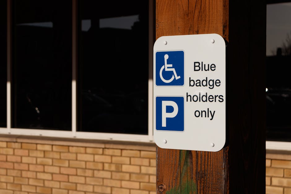A close-up image of a rectangular white parking sign mounted on a wooden post outside a building with brick exterior and large glass windows. The sign displays a blue and white wheelchair accessibility symbol accompanied by the text 'Blue badge holders only,' and a blue letter 'P' symbol. The sign indicates designated parking restrictions for mobility aid users. The area appears to be used for vehicle parking, which contains markings related to accessible parking regulations, relevant for house removals or relocation services operating in South Kensington. This visual context supports considerations for parking permits and logistical planning during furniture transport or home relocation, as managed by South Kensington Removals, especially in urban environments requiring careful management of parking permits and loading zones for efficient loading and unloading processes.