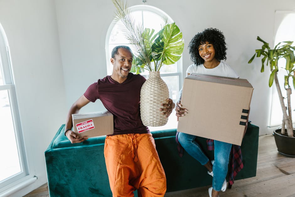 A man and a woman are inside a well-lit, modern living room with white walls and large arched windows. The man, dressed in a maroon t-shirt and orange trousers, is holding a small cardboard box marked