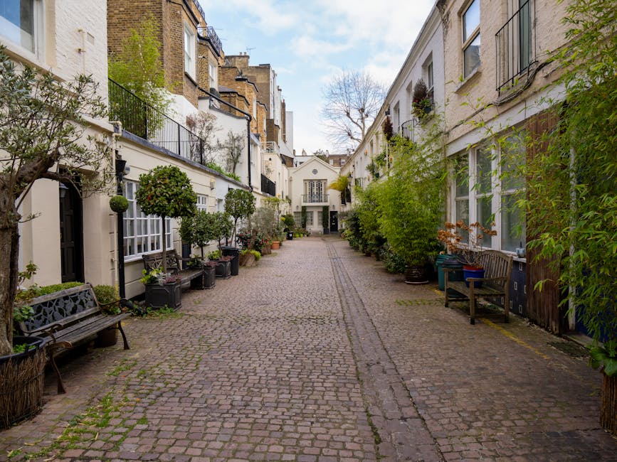 A cobblestone courtyard situated between residential buildings in South Kensington, with potted trees and plants along the sides, wooden benches, and a narrow pathway leading to a white building at the far end. The scene is captured during daylight hours with clear weather, showing some greenery and an urban setting suitable for home relocation or moving services. South Kensington Removals may utilize such spaces for loading and unloading furniture and boxes during a house move, emphasizing careful packing and transportation processes in a typical London residential area.