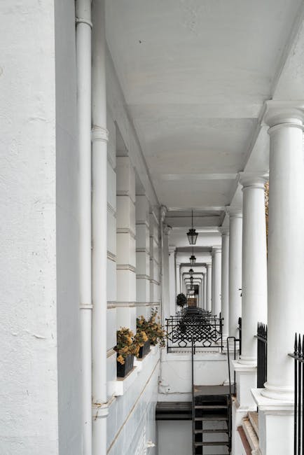 The image depicts a long, narrow residential walkway in South Kensington with white-painted exterior walls, supported by classic white columns and decorative archways. The ceiling is painted white, with traditional black outdoor lanterns hanging at intervals along the corridor. Potted plants with dried flowers are placed on window sills on the left side, while the right side features a black metal fence and a small set of wooden stairs with handrails leading down to a lower level. The pavement is made of dark stone or concrete, and the environment suggests a quiet, urban setting suitable for a home relocation project. This structural setting illustrates the tight street access typical of South Kensington properties, where careful planning is required for furniture transport and packing during removals handled by South Kensington Removals, especially for residential moves that involve delicate furniture and boxed items requiring professional loading and transport.