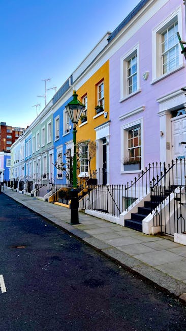 A row of colorful terraced houses in South Kensington with pastel facades painted in shades of blue, yellow, pink, and purple, situated along a narrow street with a paved pavement and a black metal railing in front of each residence. The buildings feature white-framed sash windows, some with flower boxes, and front steps leading up to doorways. A black street lamp is positioned on the sidewalk, illuminating the area during the day. In the foreground, a moving company vehicle from South Kensington Removals is partially visible, parked close to one of the houses. The van is used for home relocation and furniture transport, with its sliding door open, revealing a loading area inside. Wooden wardrobes, cardboard boxes, and wrapped furniture are seen being loaded onto or unloaded from the van by movers wearing protective gear, using trolleys and straps to facilitate the loading process. Bright daylight highlights the scene, emphasizing the neat arrangement of packing materials and the logistics involved in the removals service, reflecting typical house moving preparations in an urban setting.