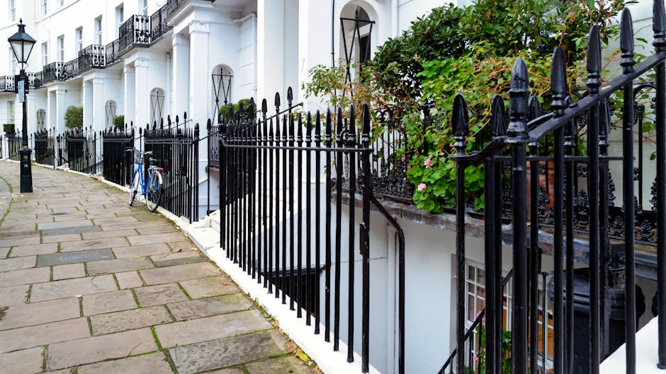 The image depicts a long, narrow residential walkway in South Kensington with white-painted exterior walls, supported by classic white columns and decorative archways. The ceiling is painted white, with traditional black outdoor lanterns hanging at intervals along the corridor. Potted plants with dried flowers are placed on window sills on the left side, while the right side features a black metal fence and a small set of wooden stairs with handrails leading down to a lower level. The pavement is made of dark stone or concrete, and the environment suggests a quiet, urban setting suitable for a home relocation project. This structural setting illustrates the tight street access typical of South Kensington properties, where careful planning is required for furniture transport and packing during removals handled by South Kensington Removals, especially for residential moves that involve delicate furniture and boxed items requiring professional loading and transport.