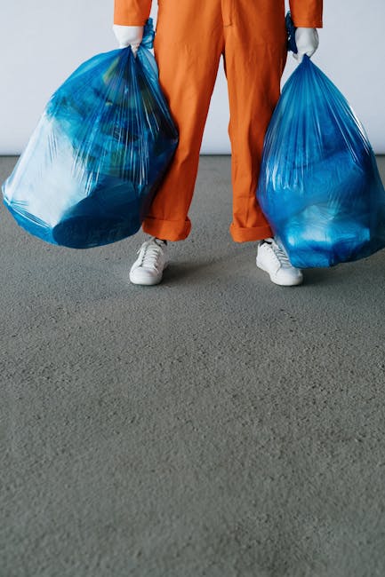 A person dressed in orange work overalls and white sneakers standing on a grey indoor surface, holding two large blue plastic rubbish bags filled with waste materials, one in each hand. The bags appea